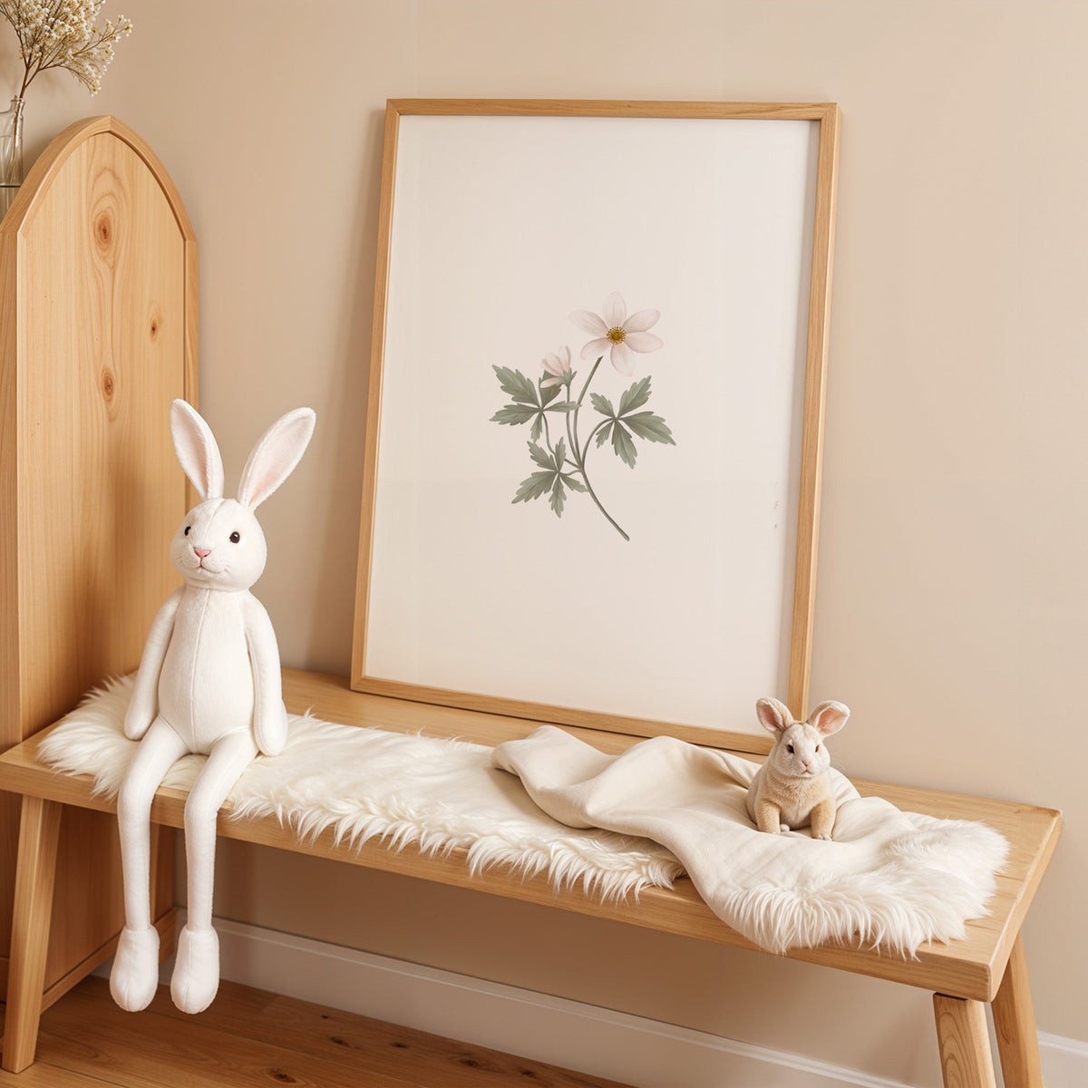 White anemone-style wildflower sprig nursery print in an oak frame on a sheepskin bench beside a white plush bunny
