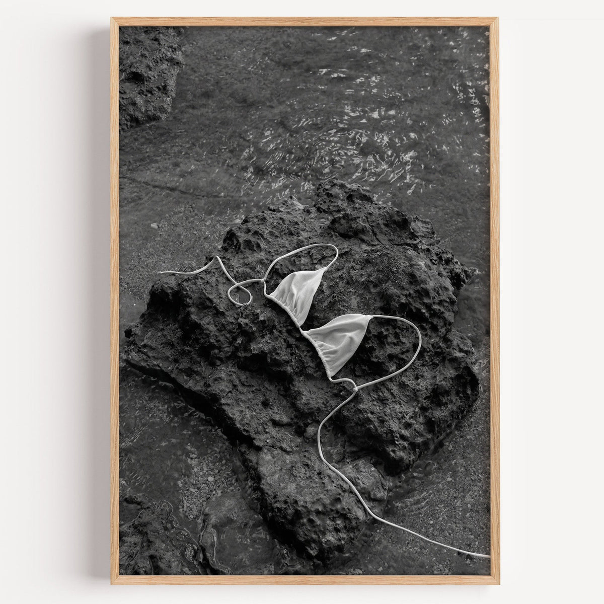 White bikini top on dark volcanic rock print in a light oak frame on a plain white wall