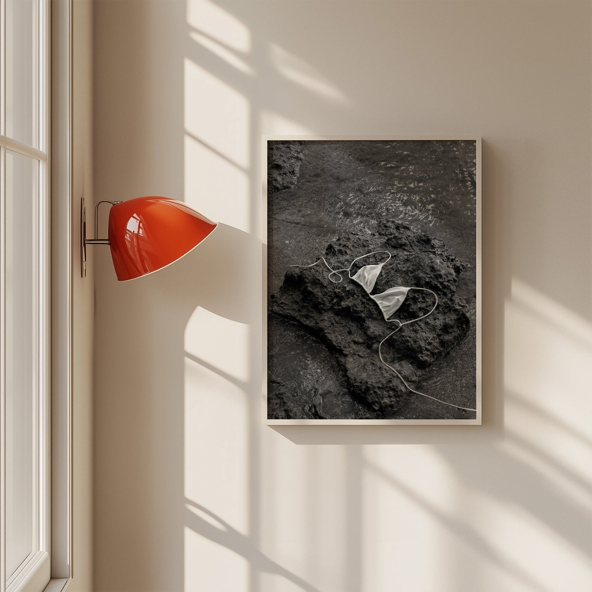 White bikini top on dark volcanic rock print in a white frame on a sunlit wall beside a red sconce