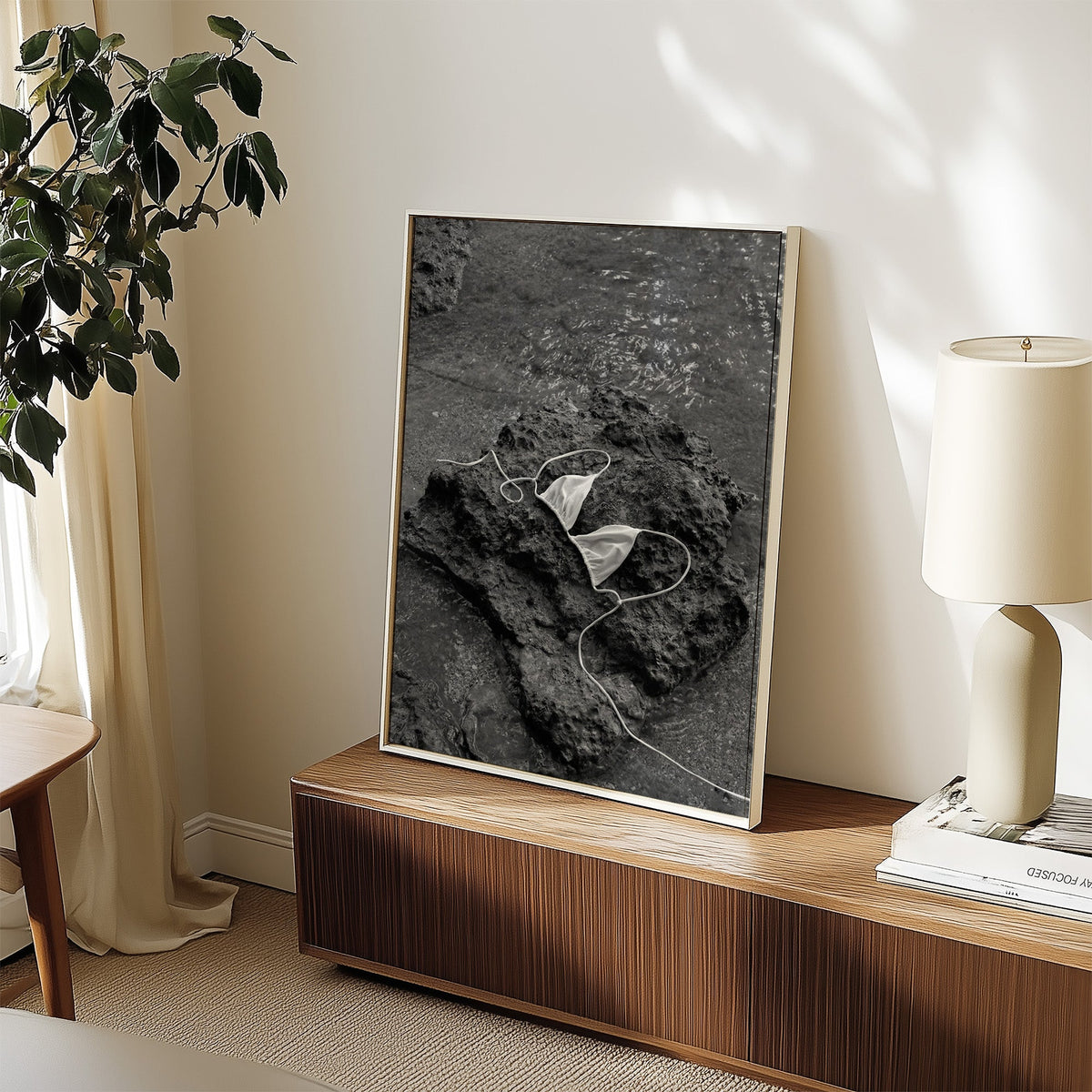 White bikini top on dark volcanic rock print in a white frame on a walnut sideboard beside a ceramic lamp