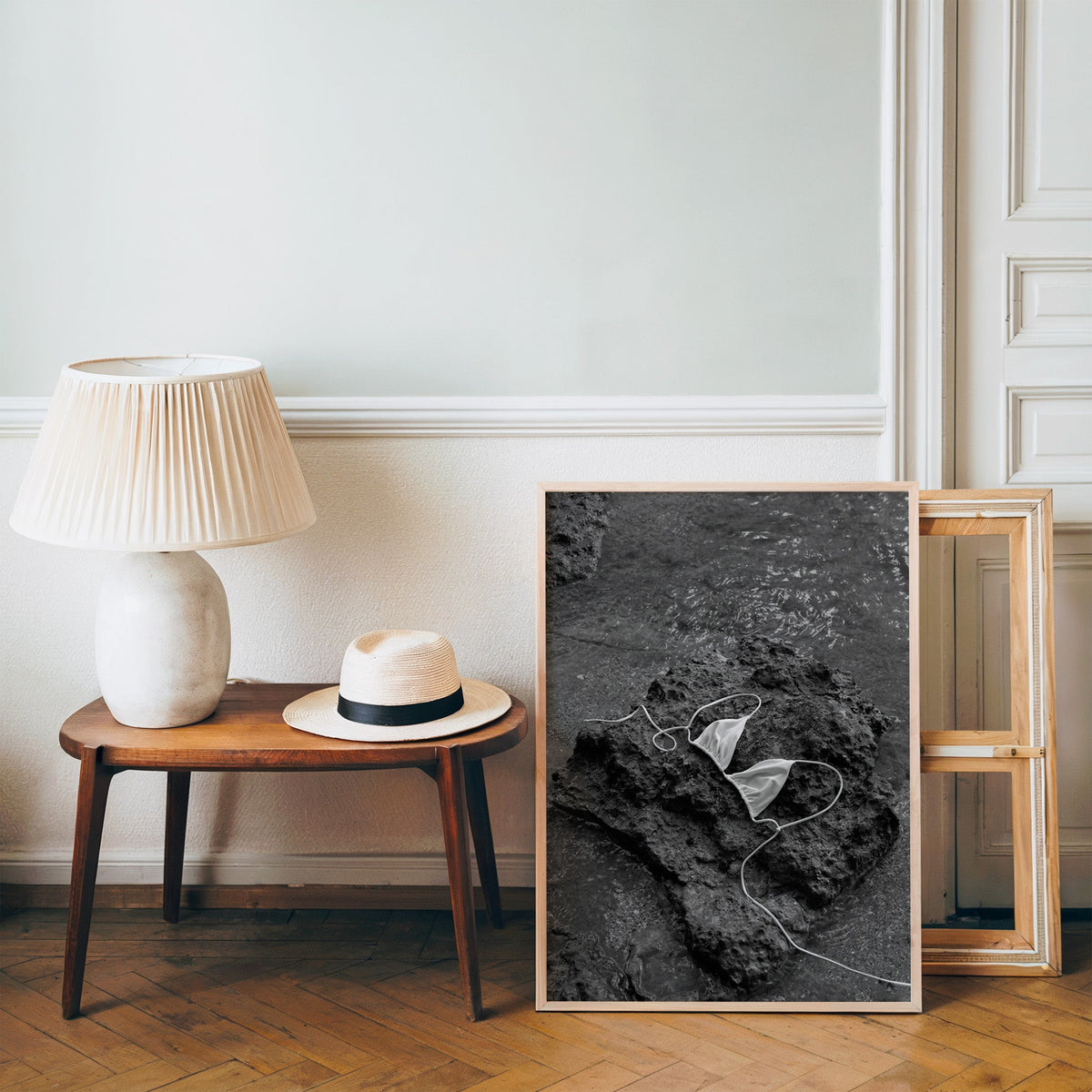 White bikini top on dark volcanic rock print in a pine frame on a wood floor beside a lamp and straw hat
