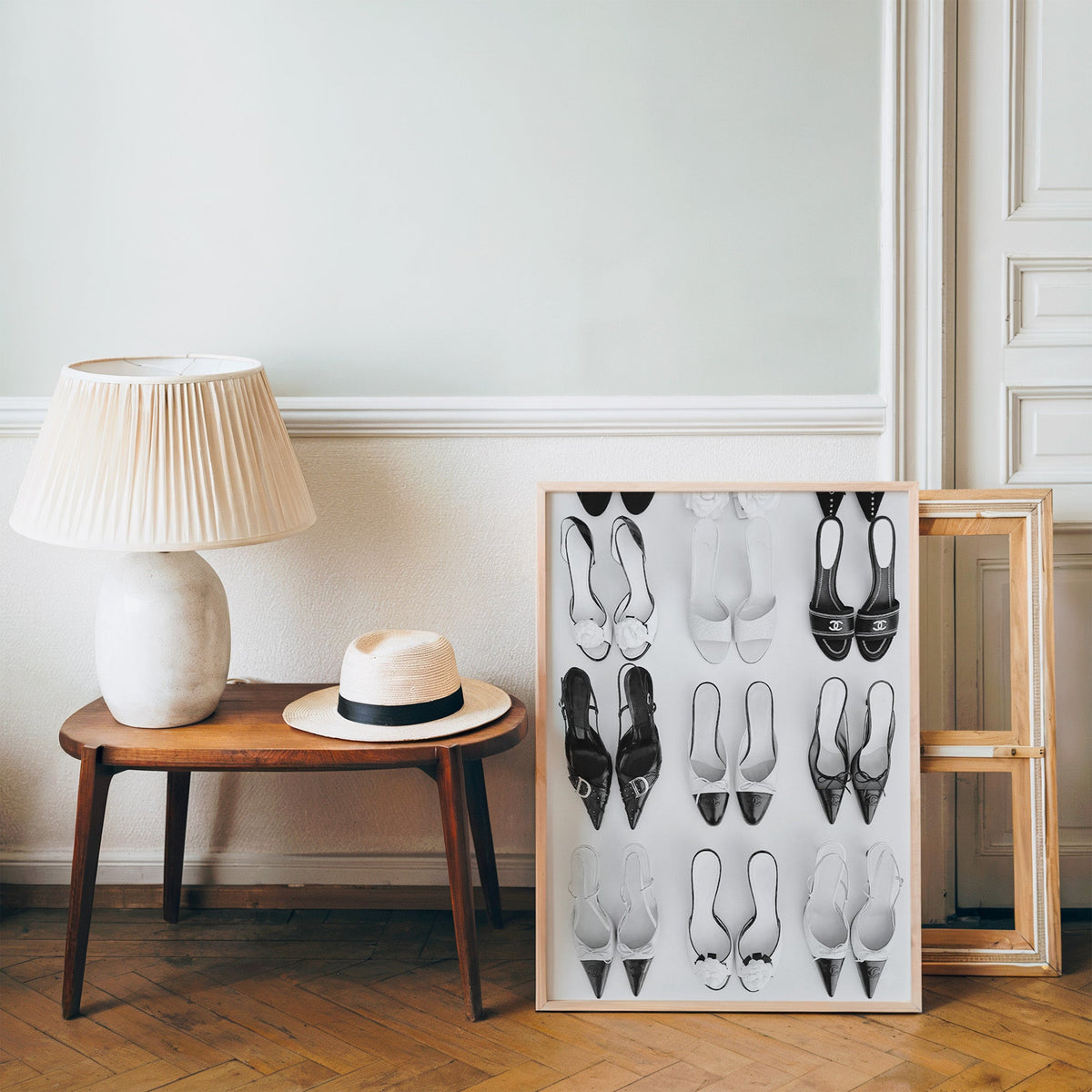 Nine pairs of mules and slingbacks in a grid print in a pine frame on a wood floor beside a lamp and straw hat