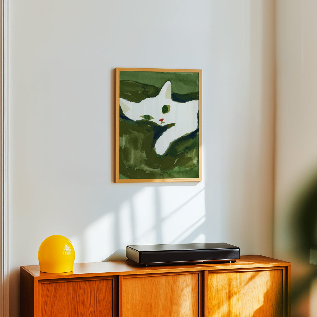 Moss green cat painting print in a light oak frame above a teak sideboard with a yellow lamp