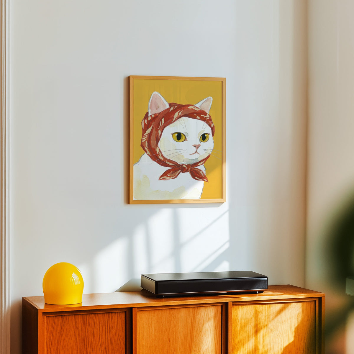 Yellow and red cat print in a light oak frame above a teak sideboard with a yellow lamp