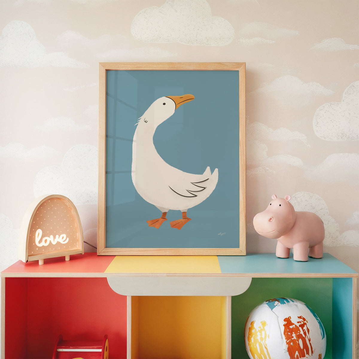 White goose on dusty blue nursery print in an oak frame on a rainbow toy shelf against cloud wallpaper