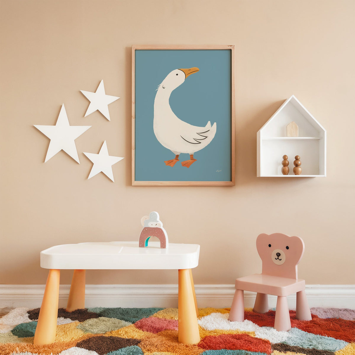 White goose on dusty blue nursery print in an oak frame on a peach wall above a kids table with wall stars