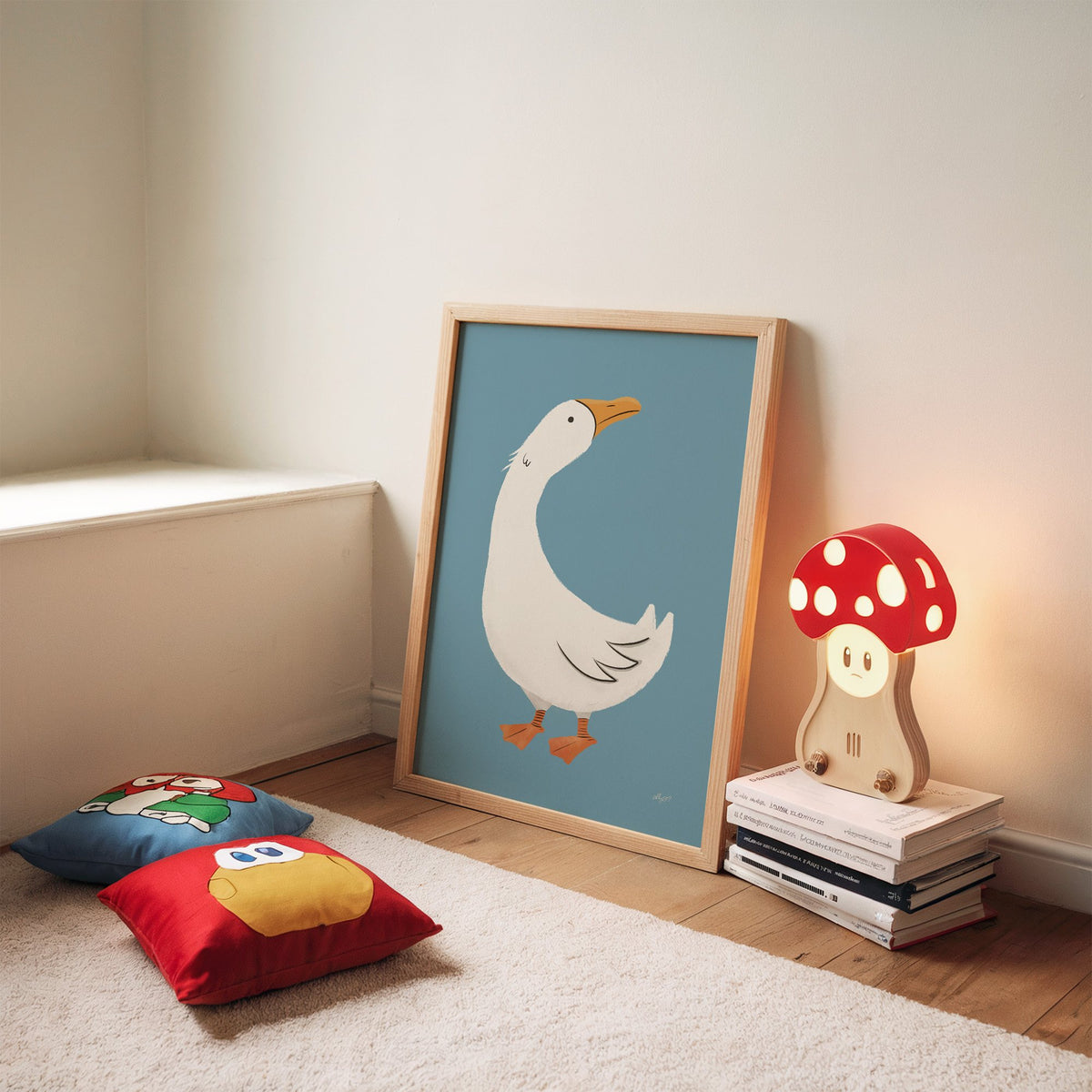 White goose on dusty blue nursery print in an oak frame leaning on a rug beside a mushroom lamp and floor cushions