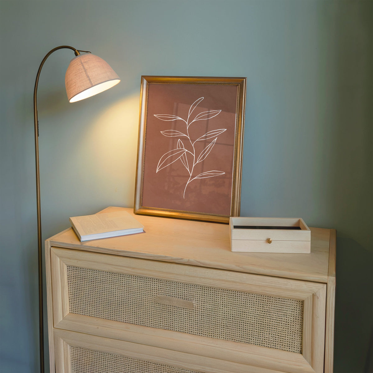 White line long leaf stem on terracotta print shown in a gold frame on a cane dresser lit by a floor lamp