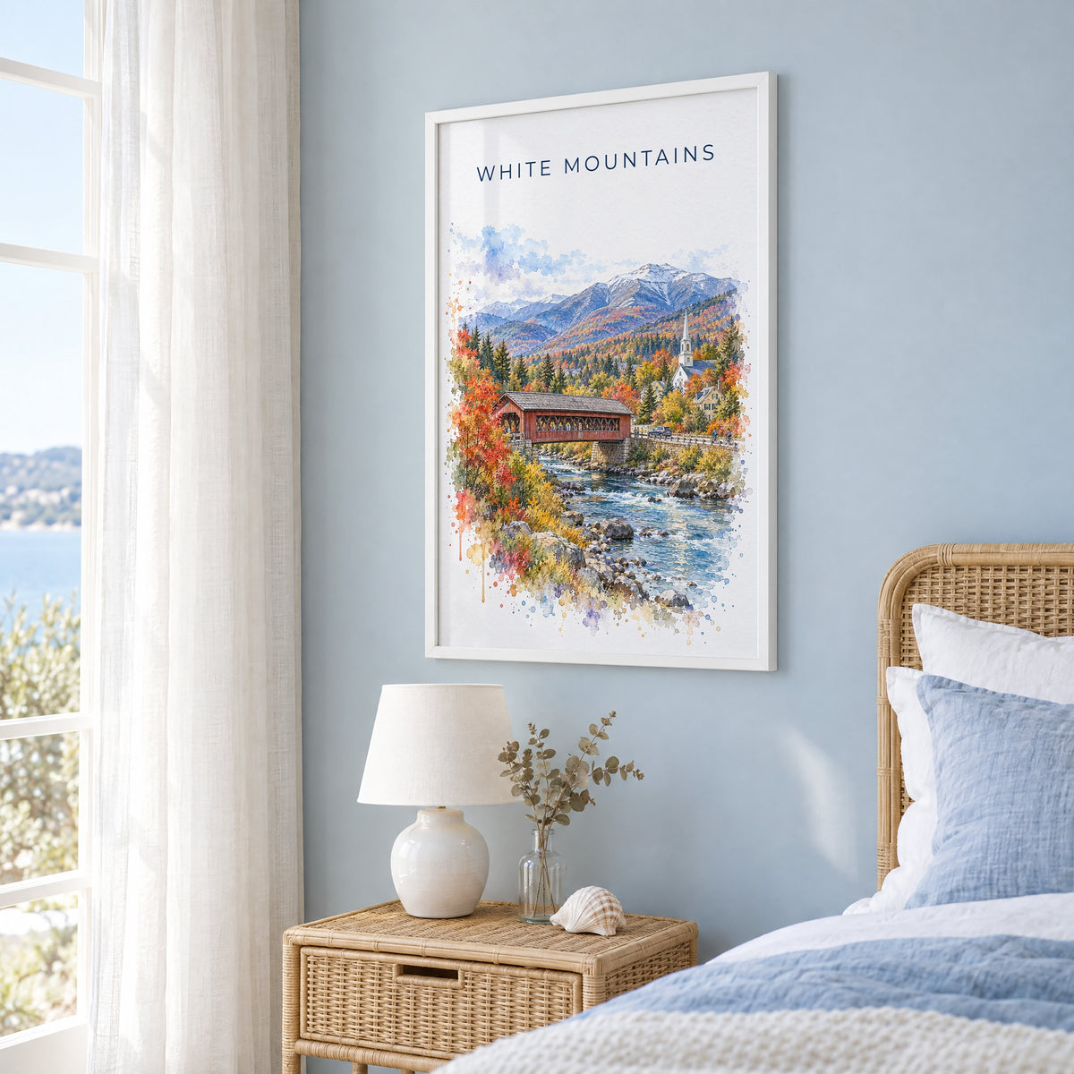 White Mountains, New Hampshire watercolor print in a white frame on a pale blue wall beside a rattan bed in a coastal bedroom