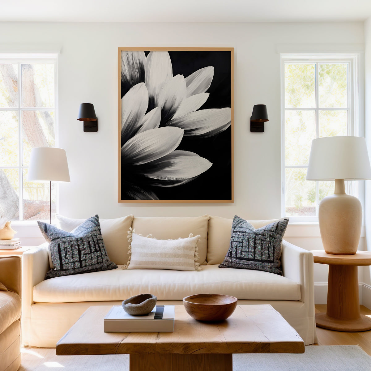 White petals on black background floral print in a light oak frame above a cream sofa between two black wall sconces
