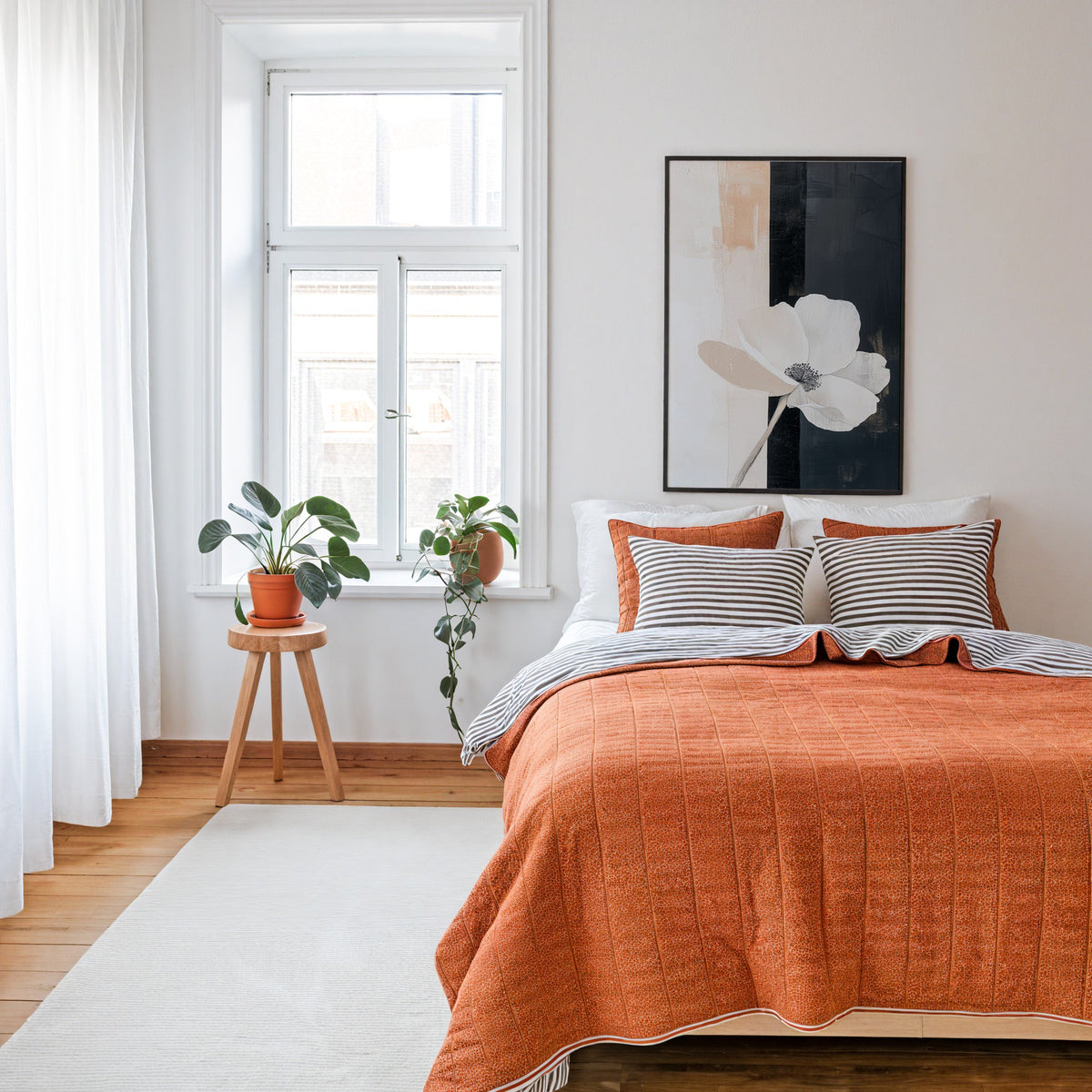 White flower on black and cream panels abstract print in a thin black frame above a bed with a rust orange quilt