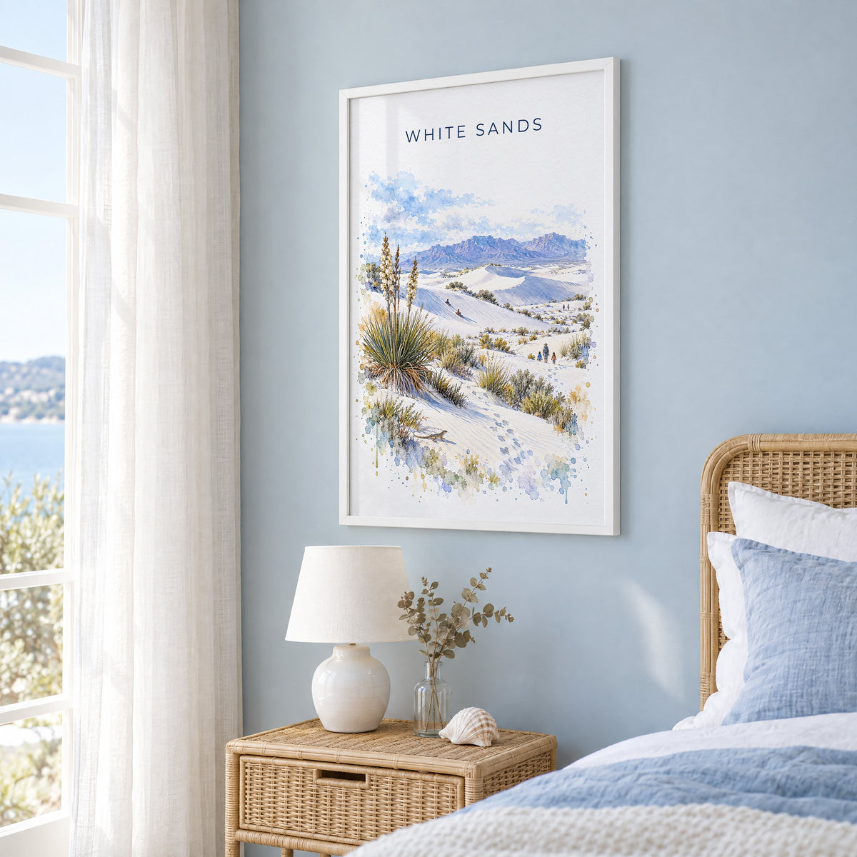 White Sands National Park watercolor print in a white frame on a pale blue wall beside a rattan bed in a coastal bedroom