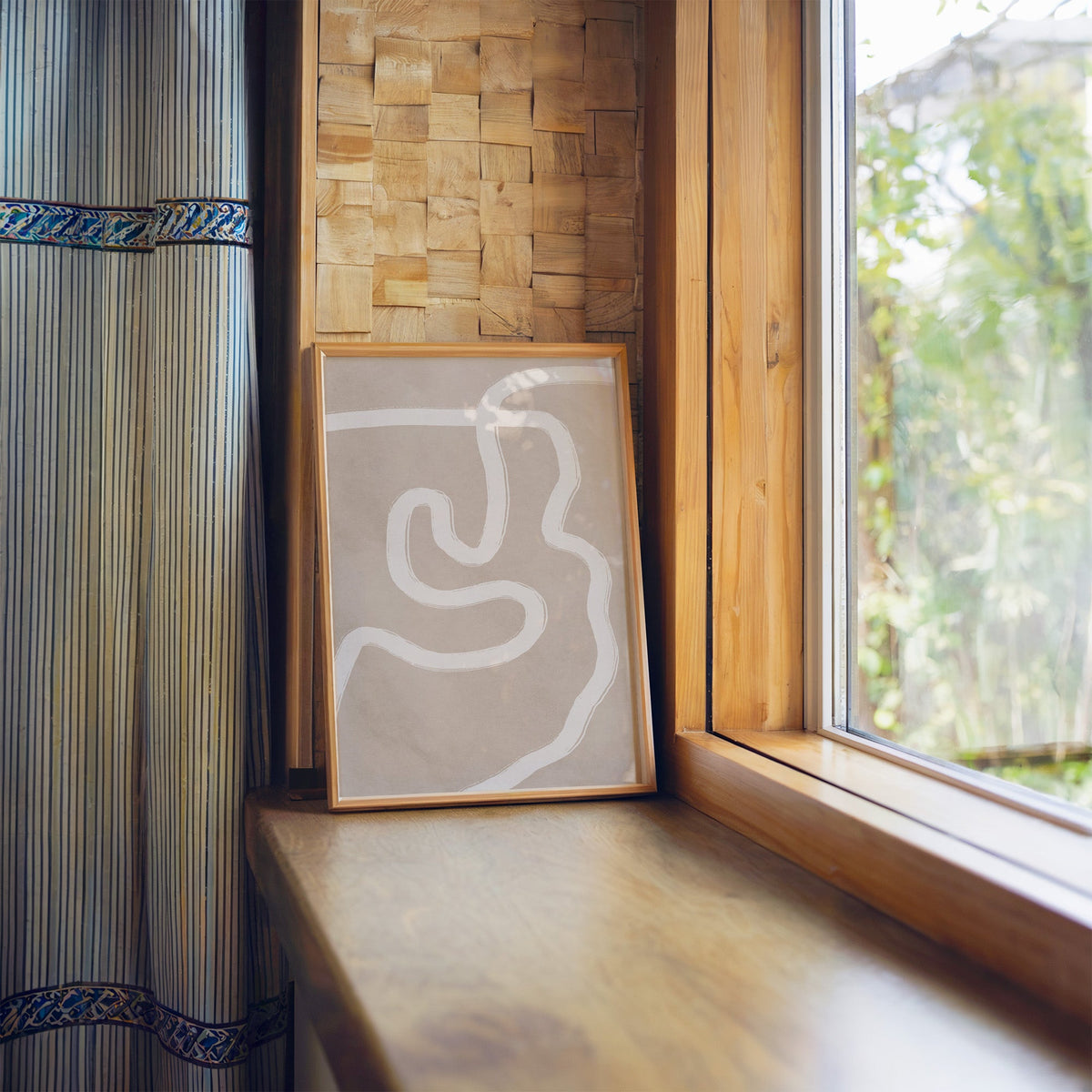 White squiggle brushstroke on taupe boho print in a light wood frame leaning on a wooden window sill by a garden view