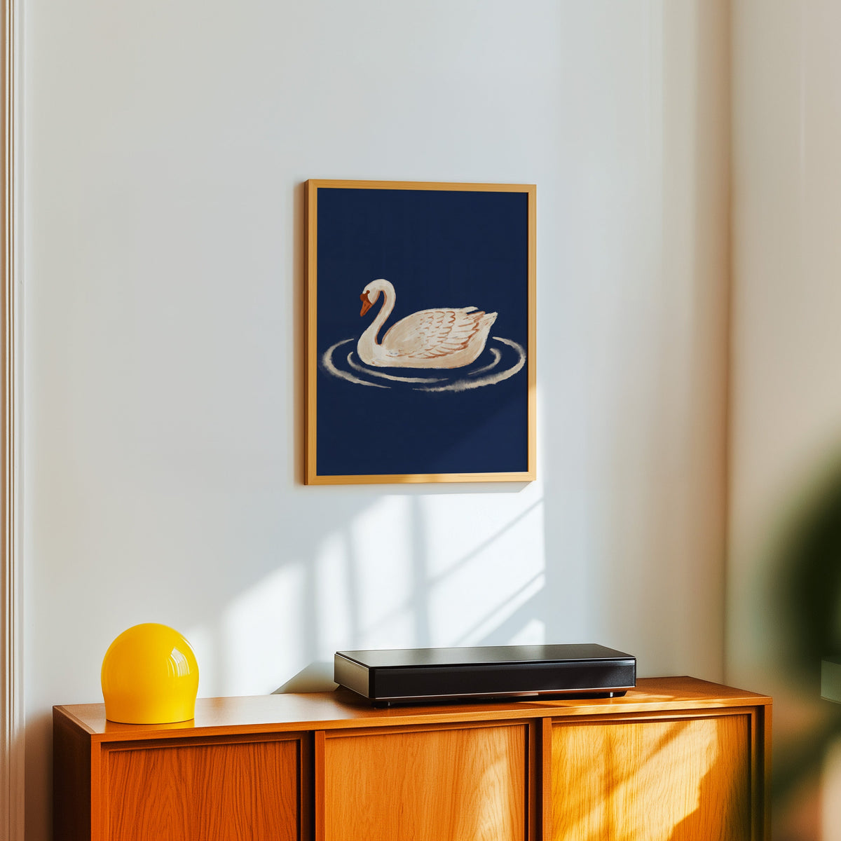 Swan and ripples print in a light oak frame above a teak sideboard with a yellow lamp
