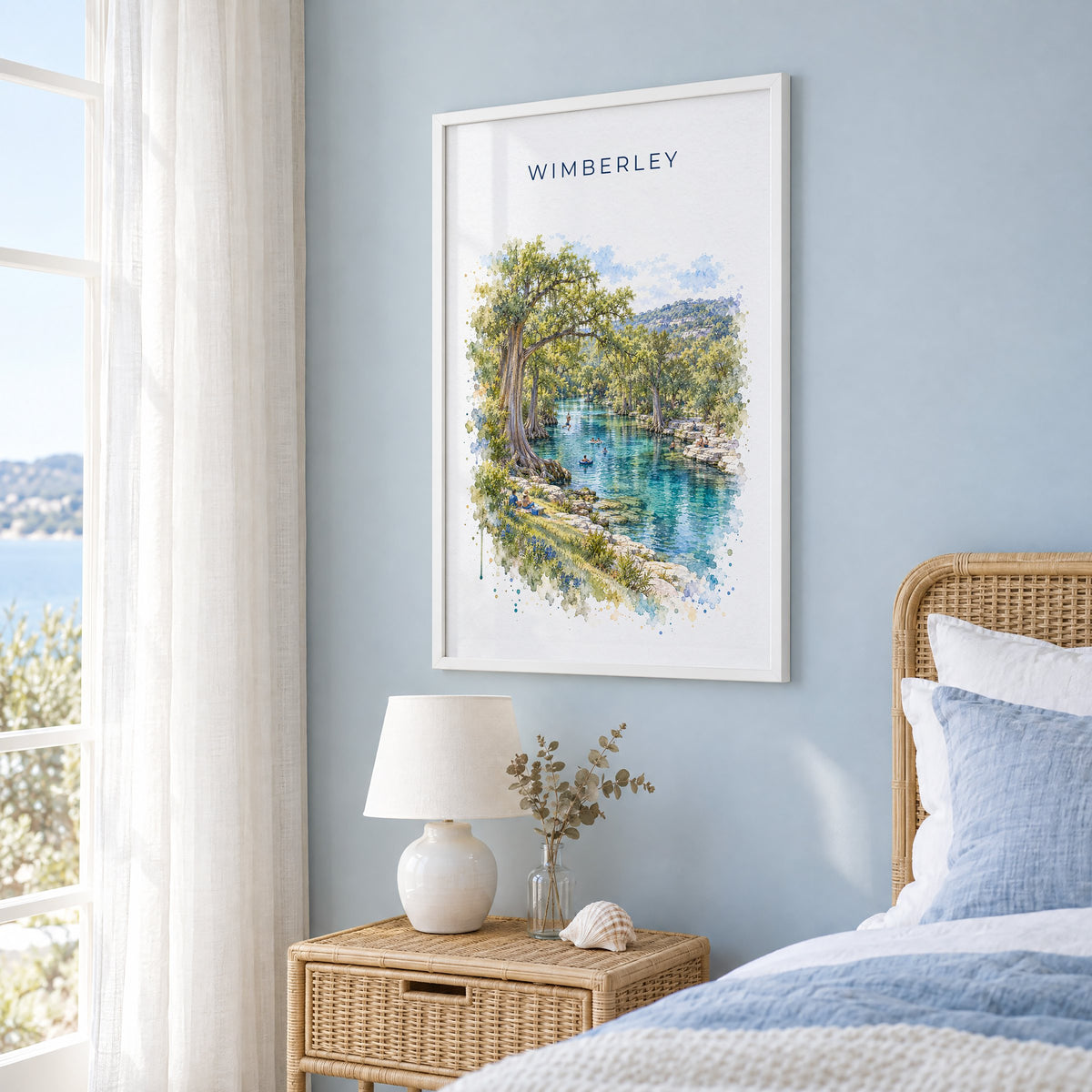 Wimberley, Texas watercolor print in a white frame on a pale blue wall beside a rattan bed in a coastal bedroom