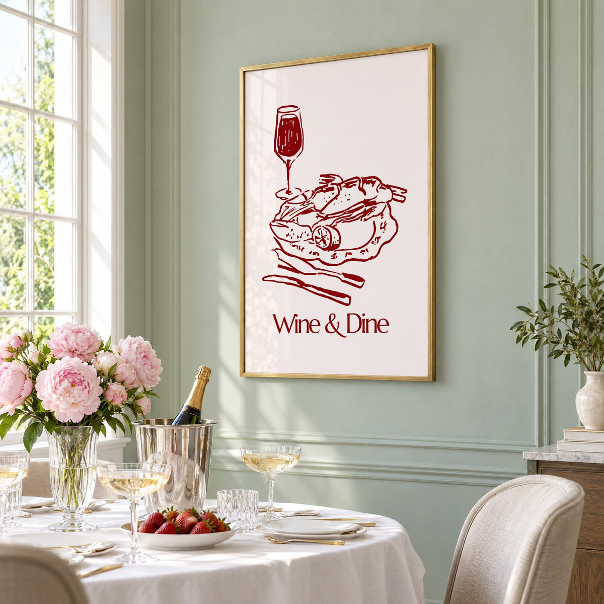 Wine & Dine roast plate burgundy sketch print in a brass frame on a sage wall above a brunch table with champagne and peonies, seen at an angle