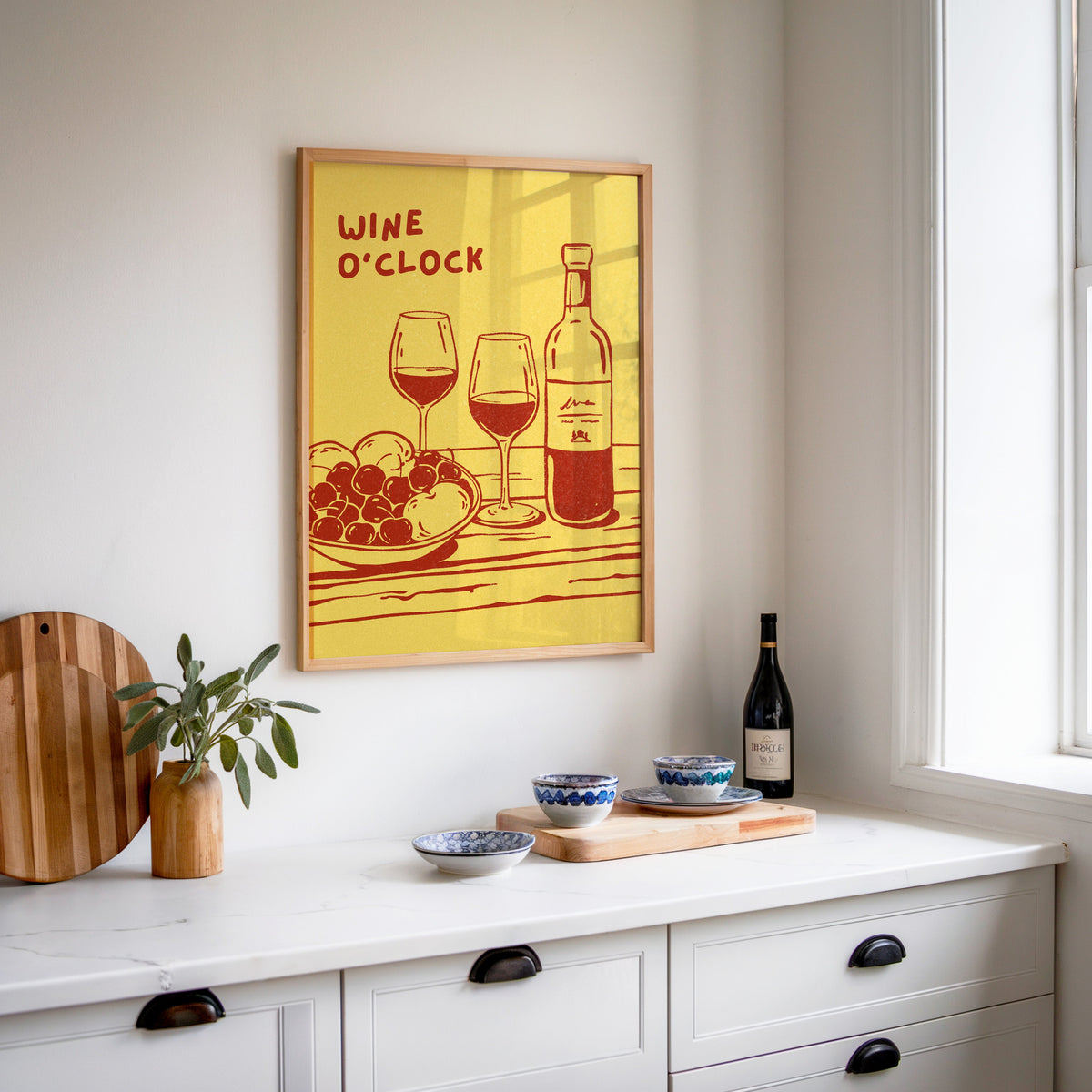 Wine O'Clock print in an oak frame above a white sideboard with a bottle of wine