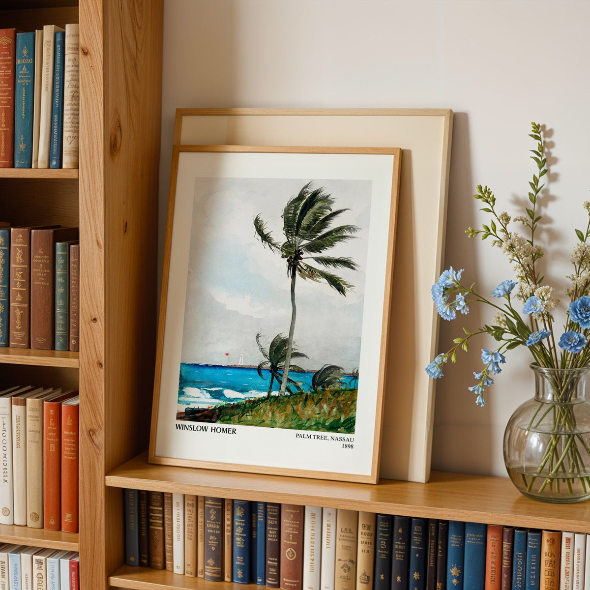 Palm tree print in an oak frame leaning on a bookshelf beside blue flowers