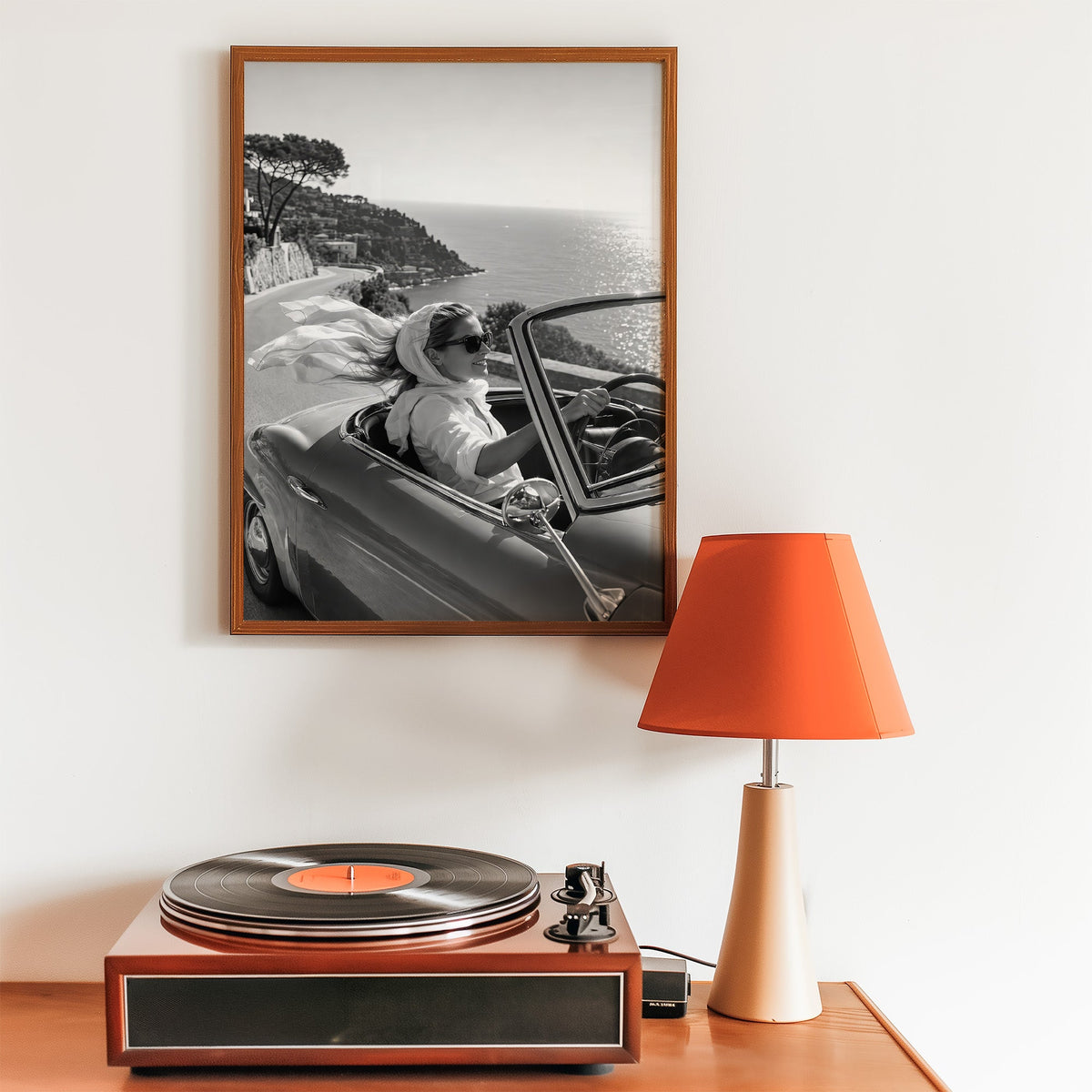Woman driving a convertible with flowing scarf print in a walnut frame above a record player and orange lamp