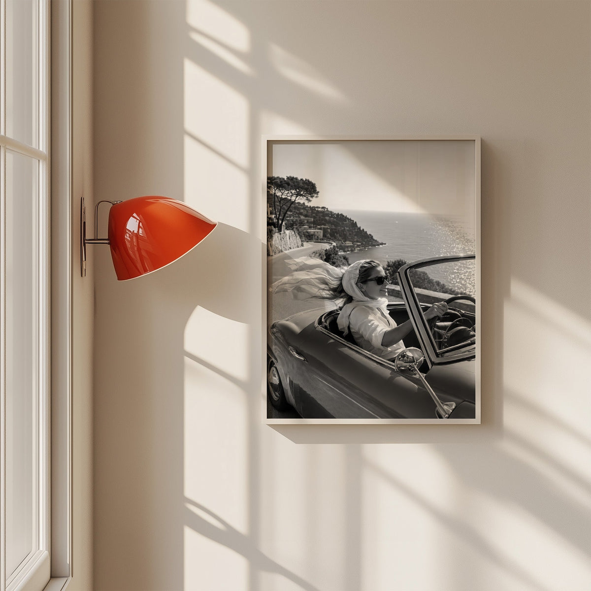 Woman driving a convertible with flowing scarf print in a white frame on a sunlit wall beside a red sconce