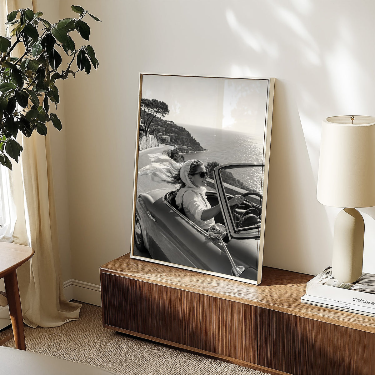 Woman driving a convertible with flowing scarf print in a white frame on a walnut sideboard beside a ceramic lamp