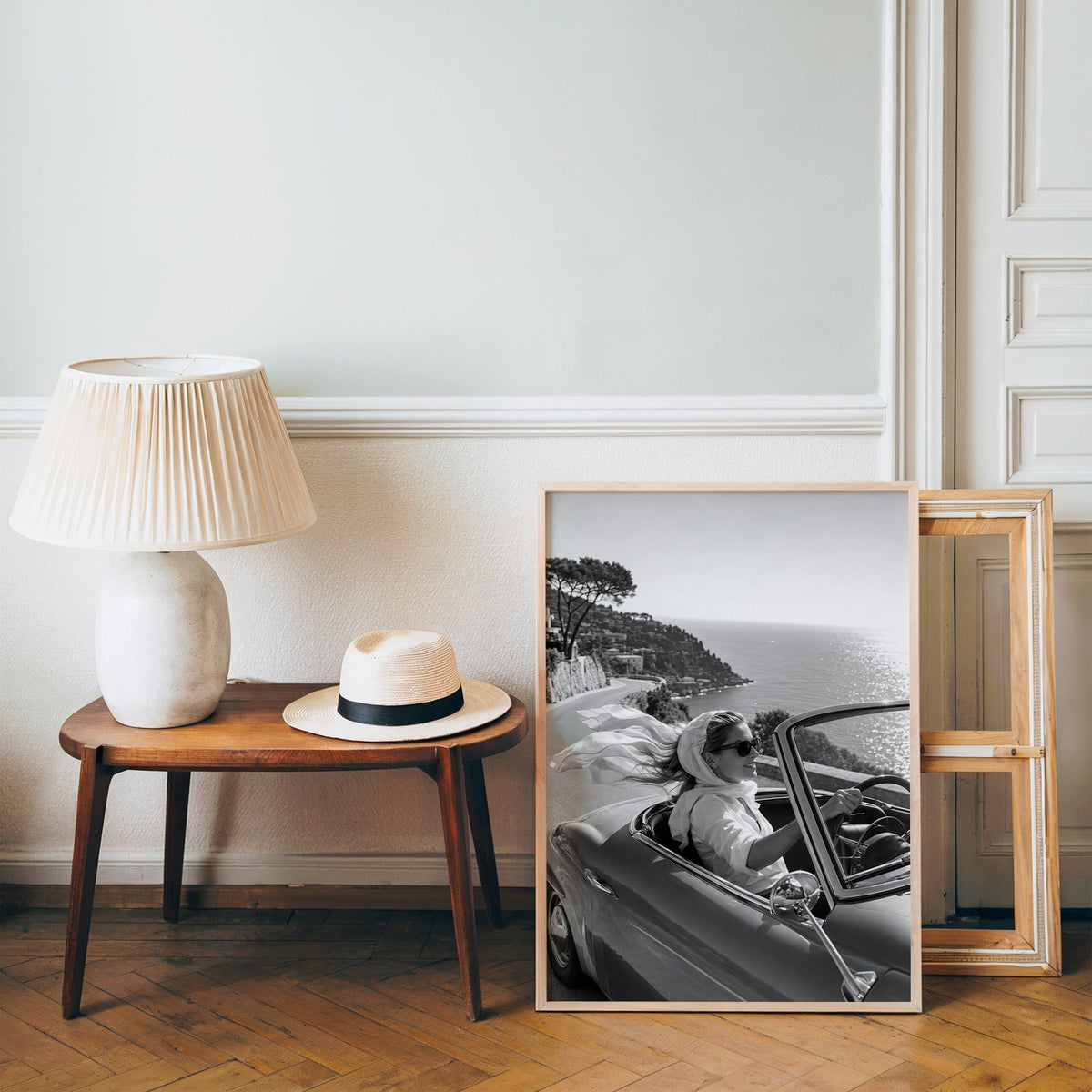 Woman driving a convertible with flowing scarf print in a pine frame on a wood floor beside a lamp and straw hat