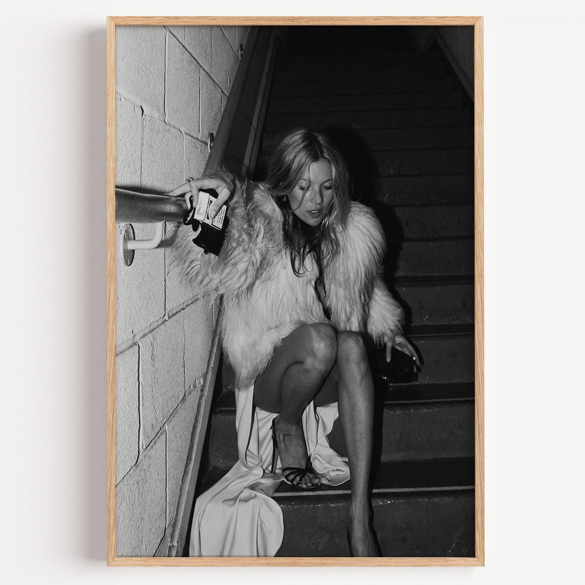 Woman in shaggy fur coat crouched on stairs print in a light oak frame on a plain white wall