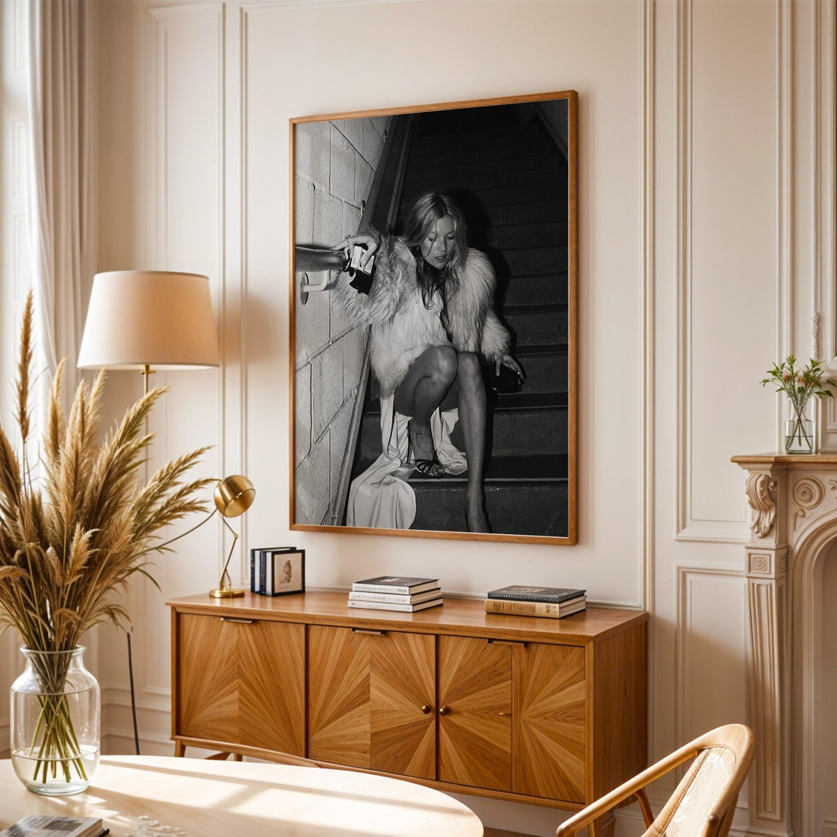 Woman in shaggy fur coat crouched on stairs print in an oak frame above a sunburst wood sideboard with pampas grass