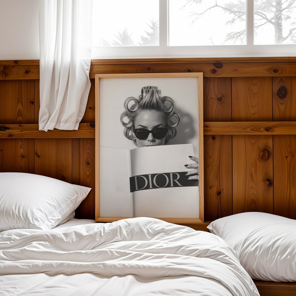 Woman in hair rollers and sunglasses behind magazine print in a pine frame leaning on a wood headboard over white bedding