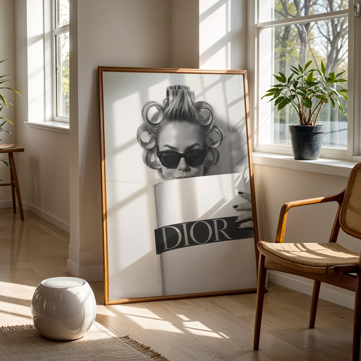 Woman in hair rollers and sunglasses behind magazine print in a walnut frame on a wood floor beside a cane chair and sunny window
