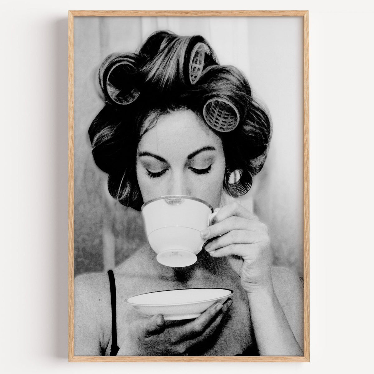 Woman in hair rollers sipping from a teacup print in a light oak frame on a plain white wall