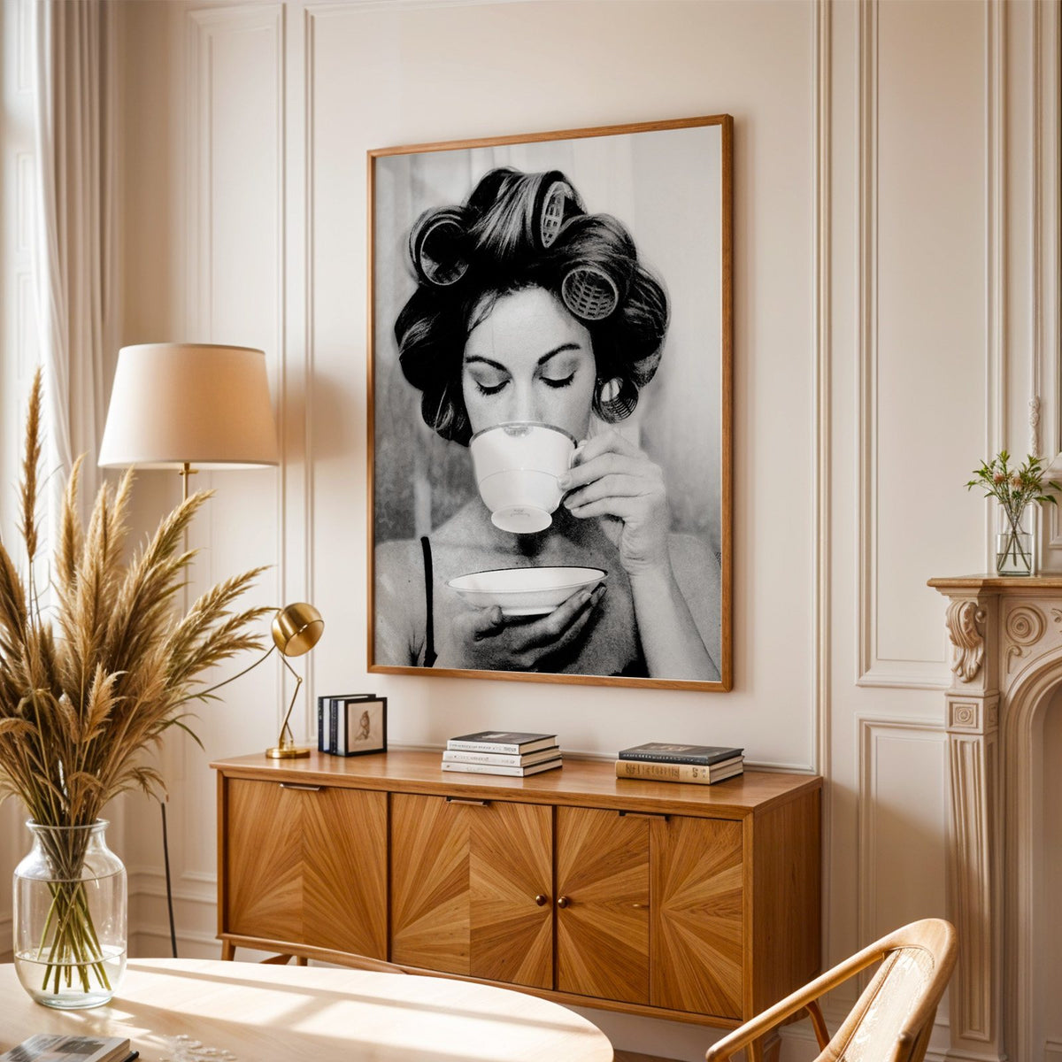 Woman in hair rollers sipping from a teacup print in an oak frame above a sunburst wood sideboard with pampas grass