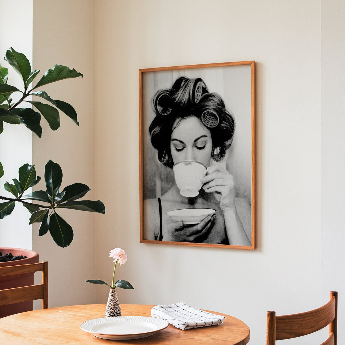 Woman in hair rollers sipping from a teacup print in a teak frame above a round wooden dining table