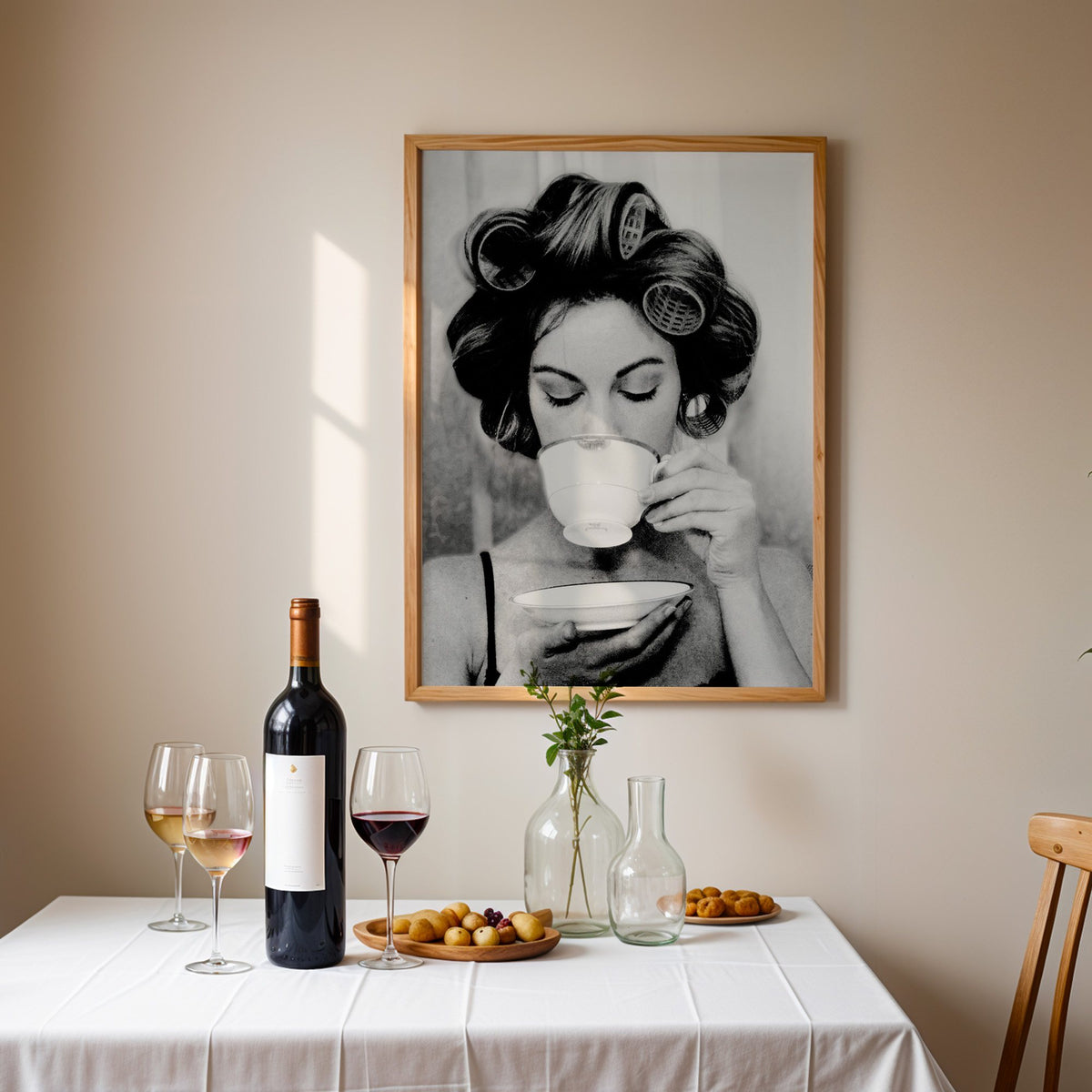 Woman in hair rollers sipping from a teacup print in an oak frame above a table set with red wine and glasses