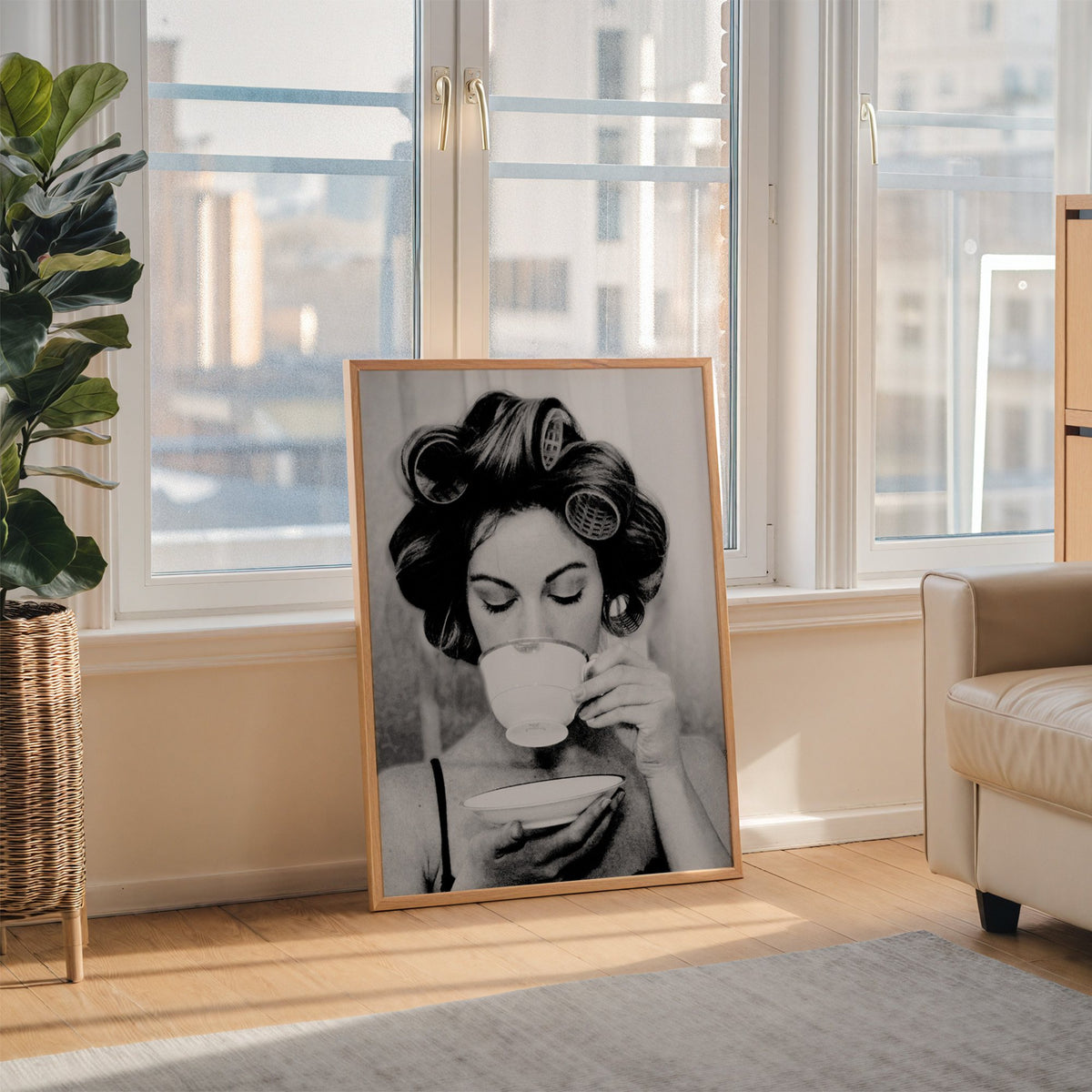 Woman in hair rollers sipping from a teacup print in an oak frame leaning on the floor beside a fiddle leaf fig