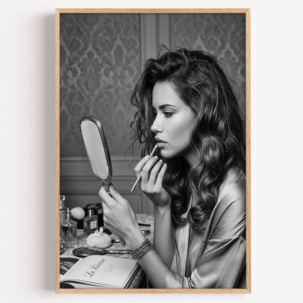 Woman lining lips with silver hand mirror print in a light oak frame on a plain white wall