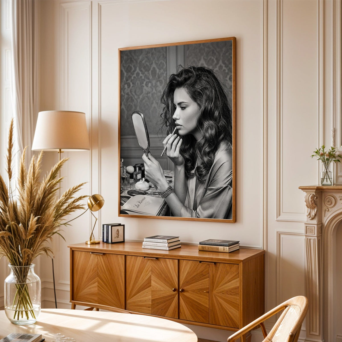 Woman lining lips with silver hand mirror print in an oak frame above a sunburst wood sideboard with pampas grass