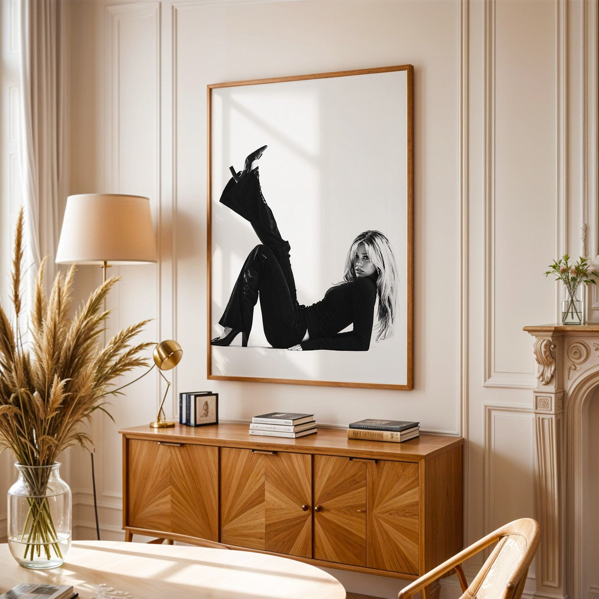 Woman in all black lying with leg raised print in an oak frame above a sunburst wood sideboard with pampas grass