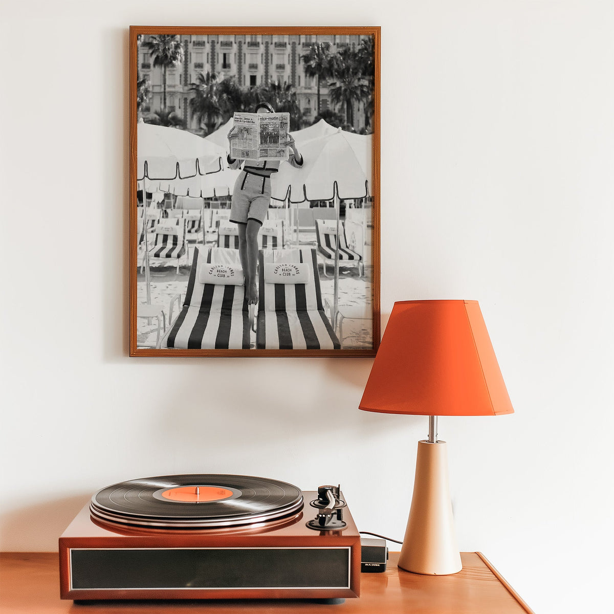 Woman in tweed suit reading a newspaper on a beach print in a walnut frame above a record player and orange lamp