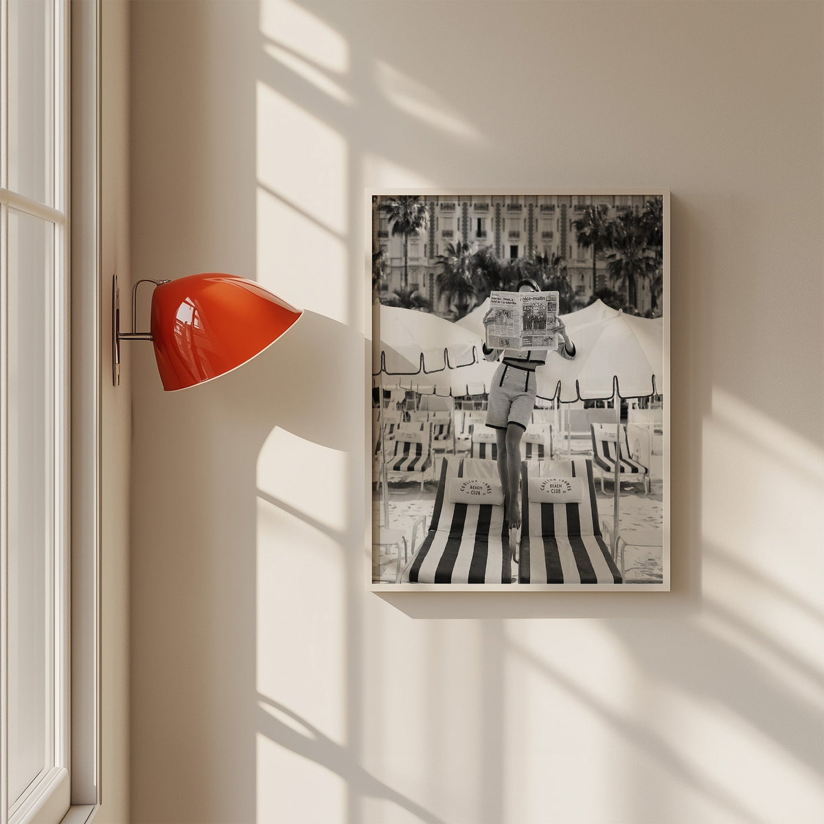 Woman in tweed suit reading a newspaper on a beach print in a white frame on a sunlit wall beside a red sconce