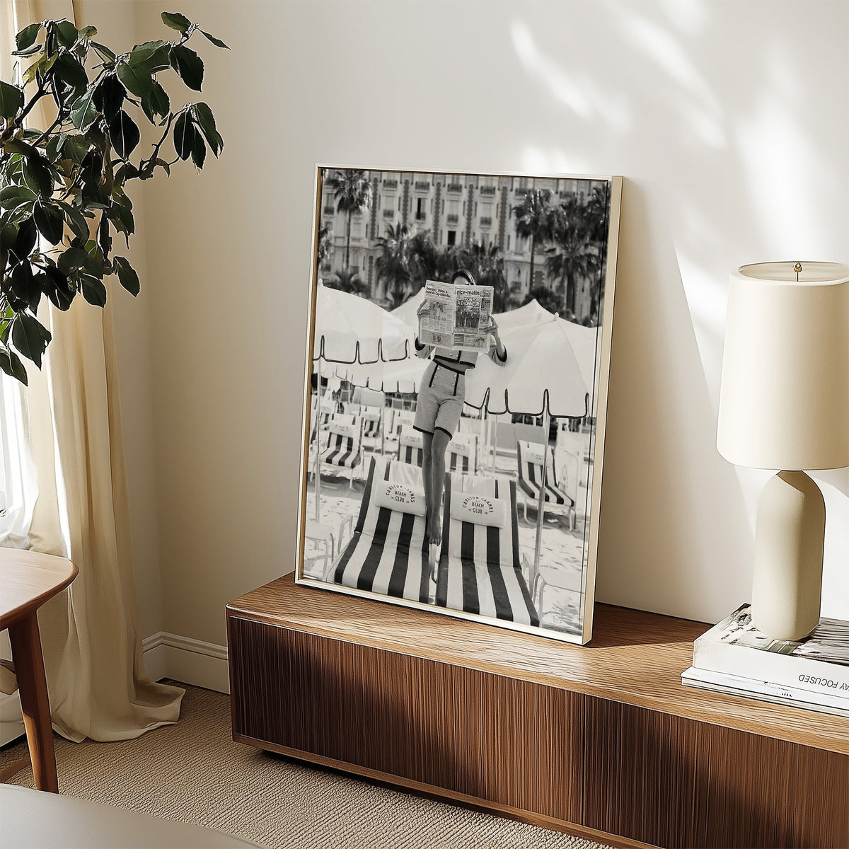 Woman in tweed suit reading a newspaper on a beach print in a white frame on a walnut sideboard beside a ceramic lamp