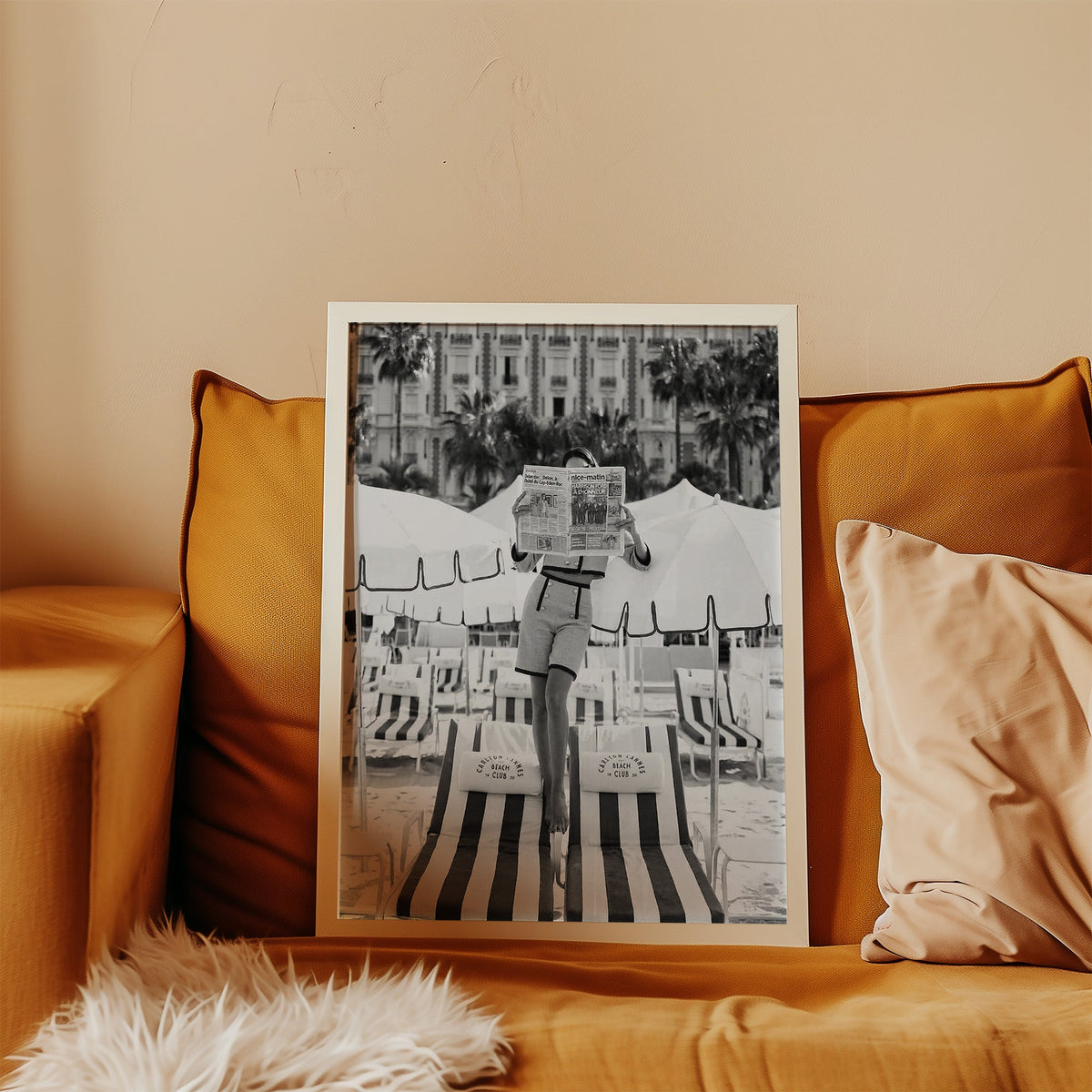 Woman in tweed suit reading a newspaper on a beach print in a white frame leaning on a mustard velvet sofa