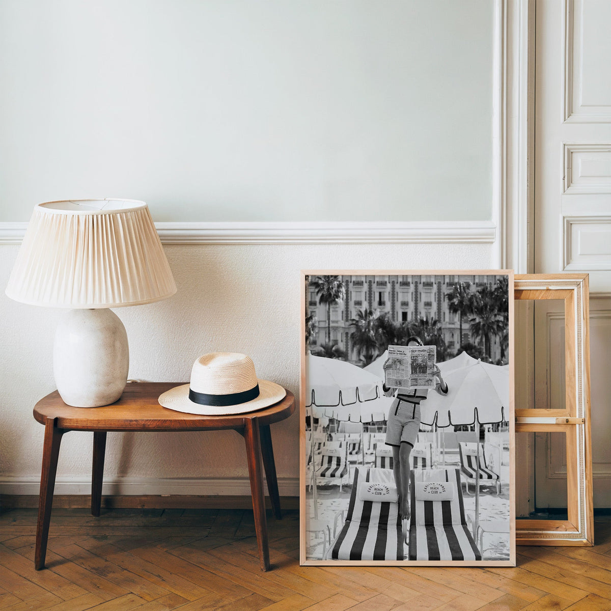 Woman in tweed suit reading a newspaper on a beach print in a pine frame on a wood floor beside a lamp and straw hat