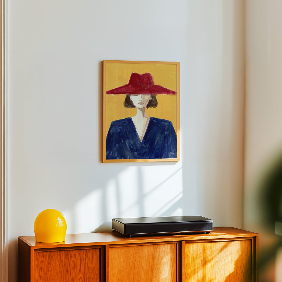 Mustard fashion illustration print in a light oak frame above a teak sideboard with a yellow lamp