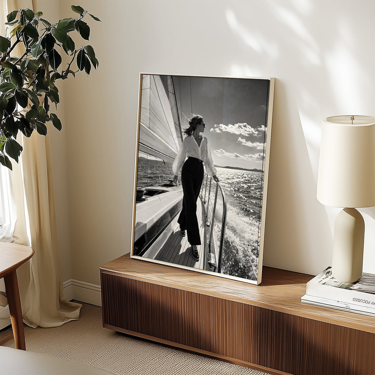 Woman in white blouse on a sailboat deck print in a white frame on a walnut sideboard beside a ceramic lamp