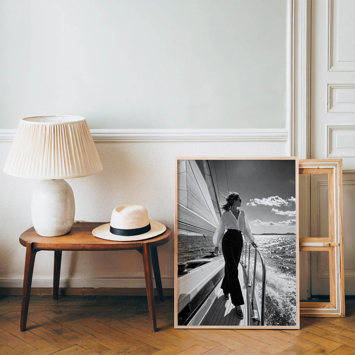 Woman in white blouse on a sailboat deck print in a pine frame on a wood floor beside a lamp and straw hat