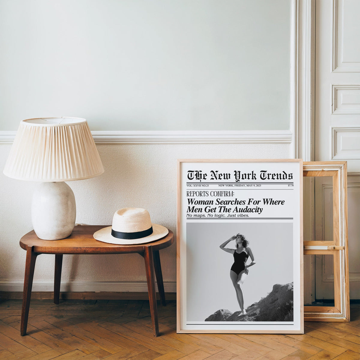 Woman on rocks shading her eyes mock newspaper print in a pine frame on a wood floor beside a lamp and straw hat