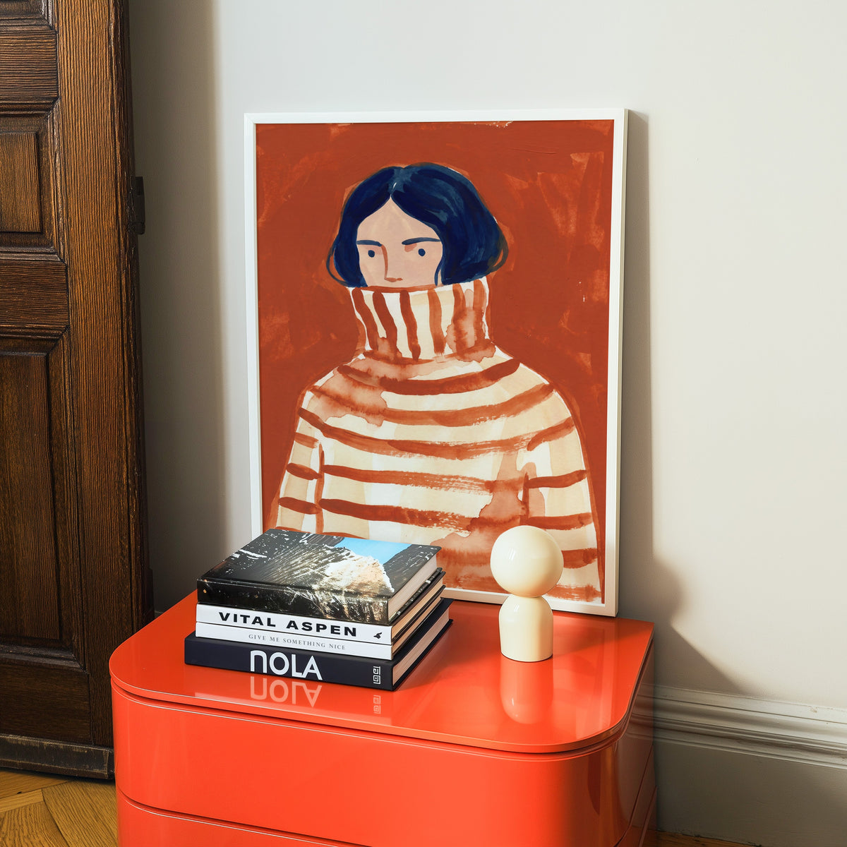 Rust watercolor portrait print in a white frame on a glossy orange cabinet with books