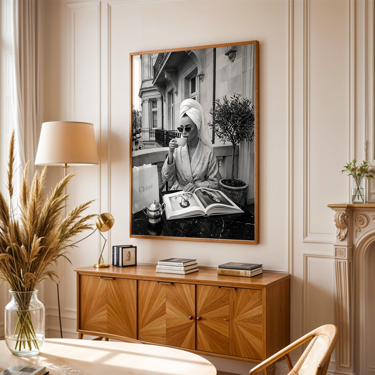 Woman in towel turban drinking coffee on balcony print in an oak frame above a sunburst wood sideboard with pampas grass
