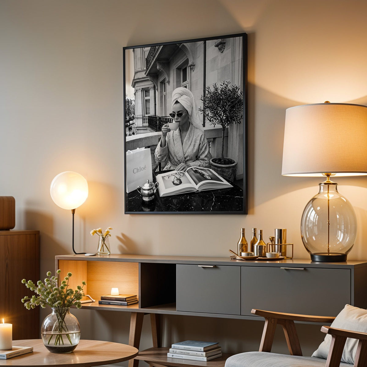 Woman in towel turban drinking coffee on balcony print in a black frame above a grey media console with a glass table lamp